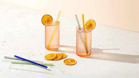Two glasses of iced citrus drinks garnished with dried orange slices, styled with colorful glass straws and a cleaning brush on a sunlit tabletop.