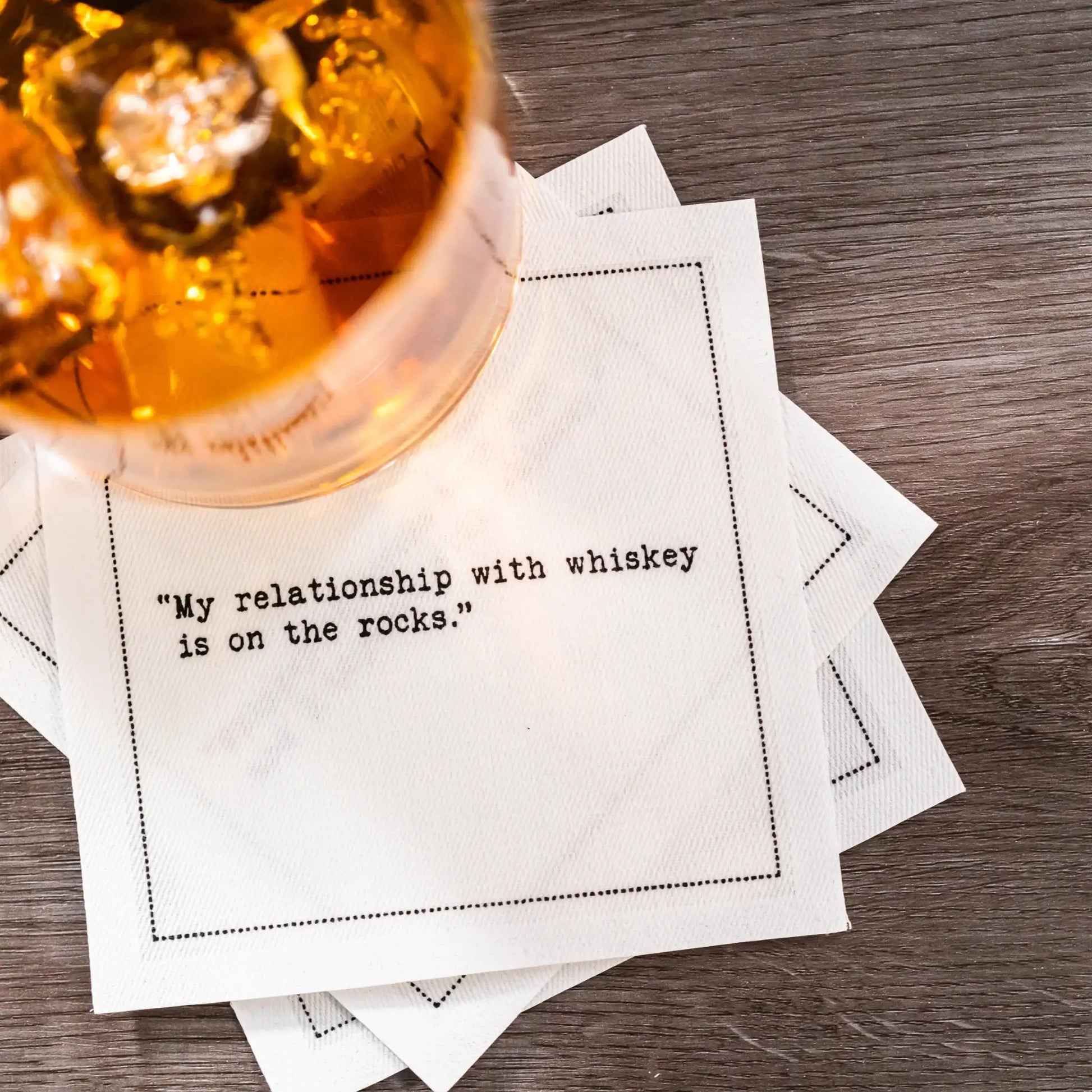 Organic cotton cocktail napkin with printed quote 'My relationship with whiskey is on the rocks.' under glass with ice, on wood table.