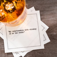 Organic cotton cocktail napkin with printed quote 'My relationship with whiskey is on the rocks.' under glass with ice, on wood table.