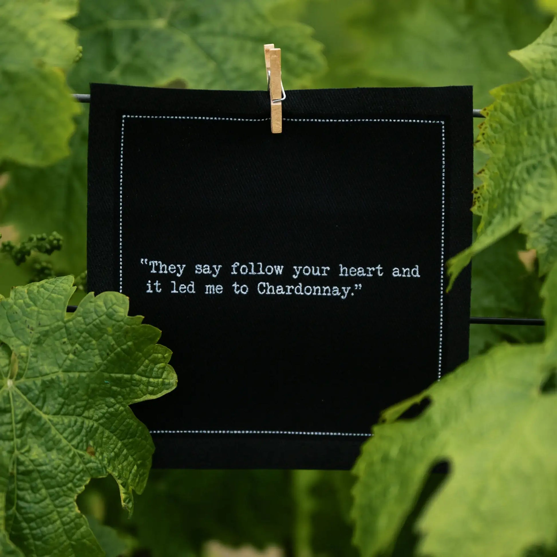 Organic cotton cocktail napkin, black, printed with the quote "They say follow your heart and it led me to Chardonnay," clipped among grapevine leaves, from Five Star Napkins' Wine quote collection