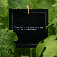 Organic cotton cocktail napkin, black, printed with the quote "They say follow your heart and it led me to Chardonnay," clipped among grapevine leaves, from Five Star Napkins' Wine quote collection