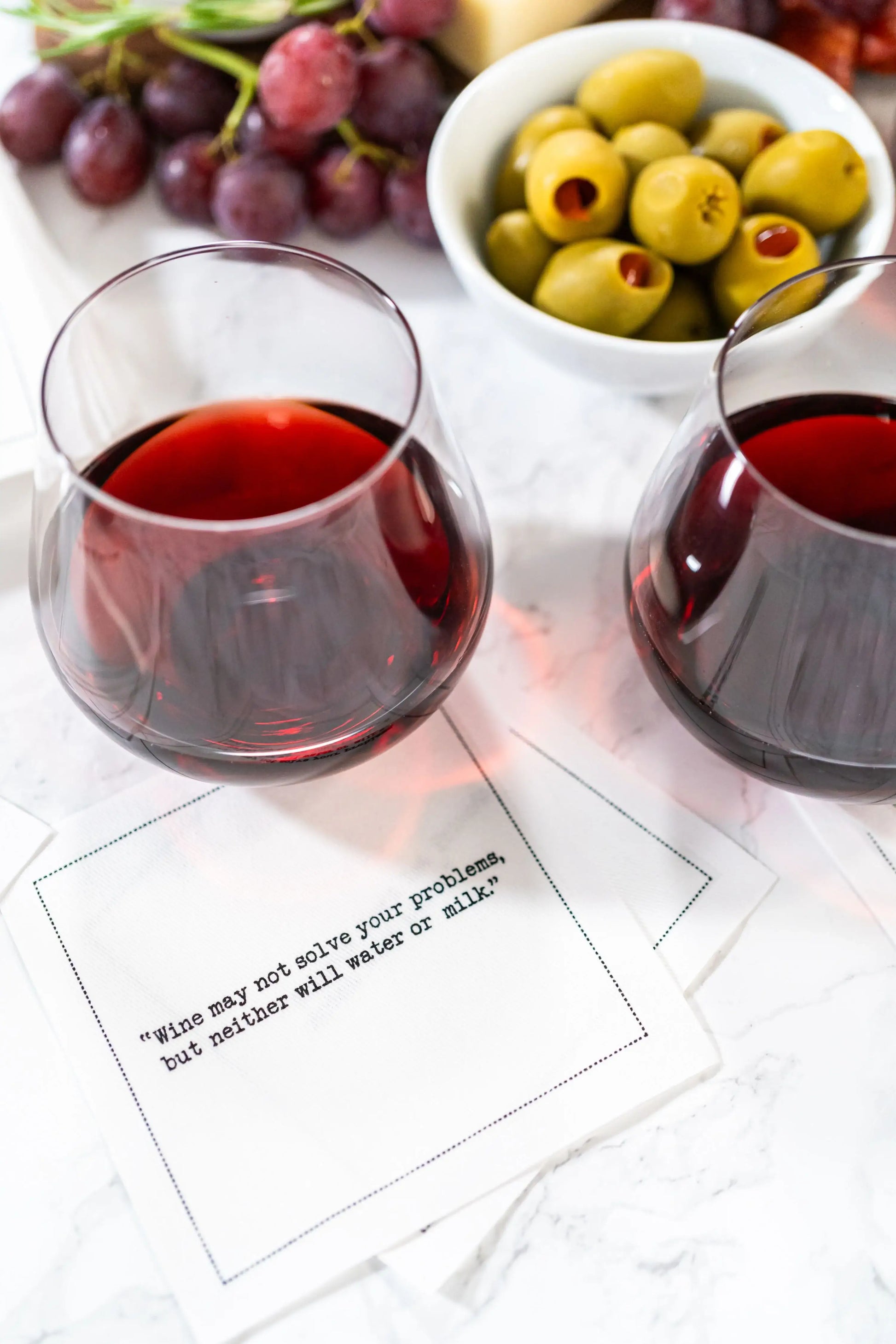 Organic cotton cocktail napkin printed with the quote "Wine may not solve your problems, but neither will water or milk," styled with red wine, grapes, olives, and cheese on a marble table, from Five Star Napkins' Wine quote collection