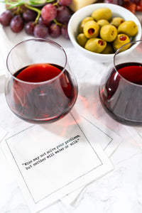 Organic cotton cocktail napkin printed with the quote "Wine may not solve your problems, but neither will water or milk," styled with red wine, grapes, olives, and cheese on a marble table, from Five Star Napkins' Wine quote collection