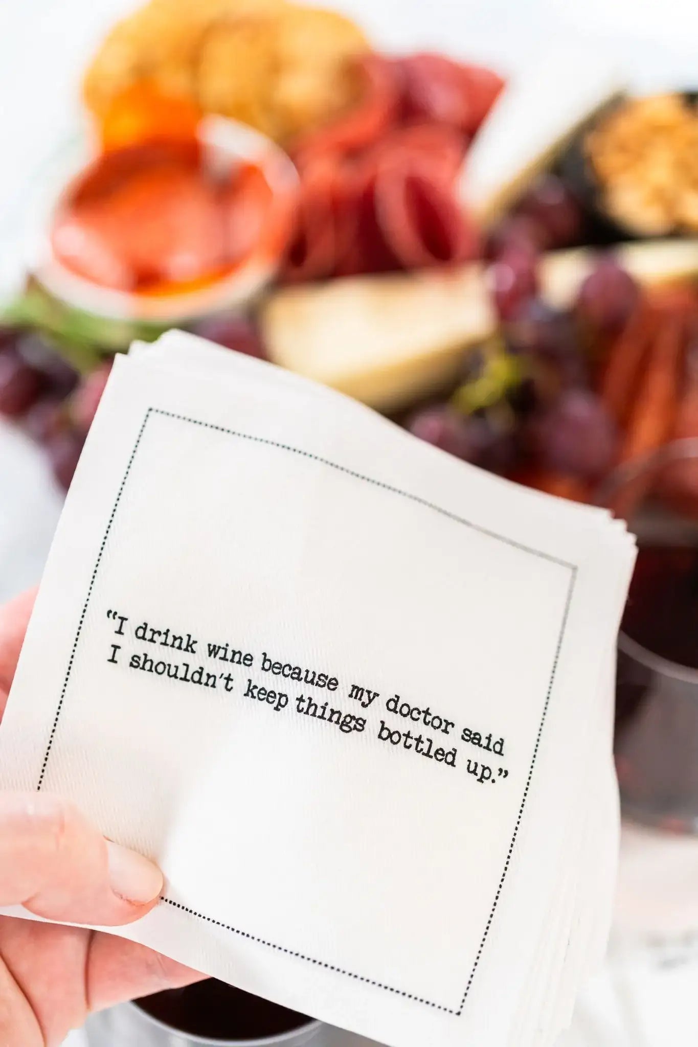 Organic cotton cocktail napkin printed with the quote "I drink wine because my doctor said I shouldn't keep things bottled up," held near a charcuterie board, from Five Star Napkins' Wine quote collection