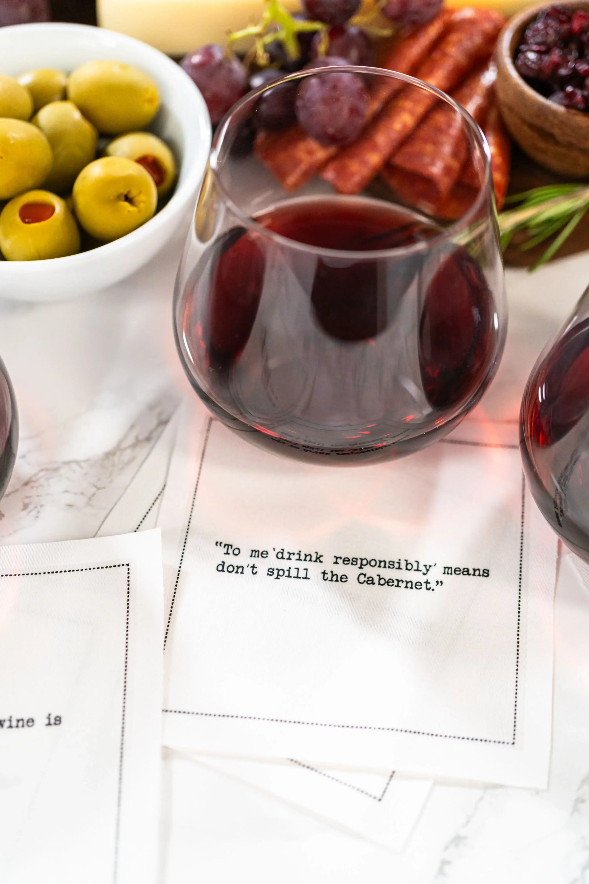 Organic cotton cocktail napkin printed with the quote "To me 'drink responsibly' means don't spill the Cabernet," styled with wine glasses, olives, and charcuterie on a marble table, from Five Star Napkins' Wine quote collection