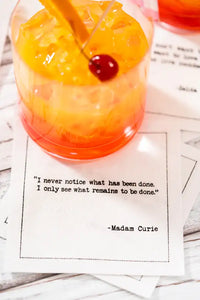 Organic cotton cocktail napkin with quote 'I never notice what has been done. I only see what remains to be done.' by Madam Curie, under a cherry-garnished drink at a cocktail party setting.