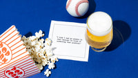 Organic cotton cocktail napkin printed with the quote “I wish I had an answer to that because I’m tired of answering that question,” – Yogi Berra, styled with popcorn, a baseball, and a beer, from Five Star Napkins’ Yogi Berra quote collection.