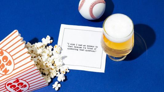 Organic cotton cocktail napkin printed with the quote “I wish I had an answer to that because I’m tired of answering that question,” – Yogi Berra, styled with popcorn, a baseball, and a beer, from Five Star Napkins’ Yogi Berra quote collection.