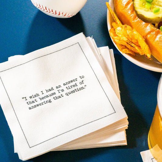 Organic cotton cocktail napkin printed with the quote “I wish I had an answer to that because I’m tired of answering that question,” – Yogi Berra, styled with a hot dog, chips, and a beer, from Five Star Napkins’ Yogi Berra quote collection.