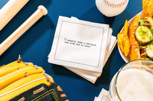 Organic cotton cocktail napkin printed with the quote “I usually take a two-hour nap from one to four,” – Yogi Berra, styled with a baseball bat, glove, and hot dog, from Five Star Napkins’ Yogi Berra quote collection.