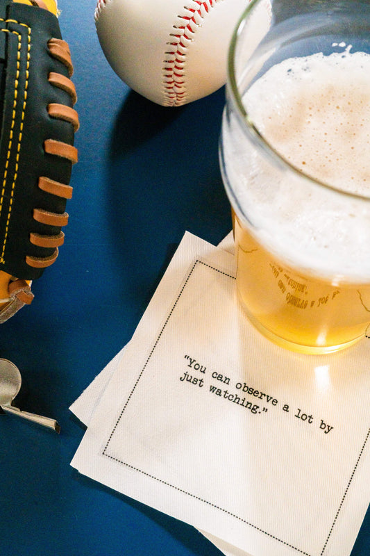 Organic cotton cocktail napkin printed with the quote “You can observe a lot by just watching,” – Yogi Berra, styled with a baseball glove and a beer, from Five Star Napkins’ Yogi Berra quote collection.
