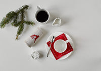Table setting with red organic cotton napkin, linen organic cotton napkin with reindeer design, coffee cup, sugar, and pine branch