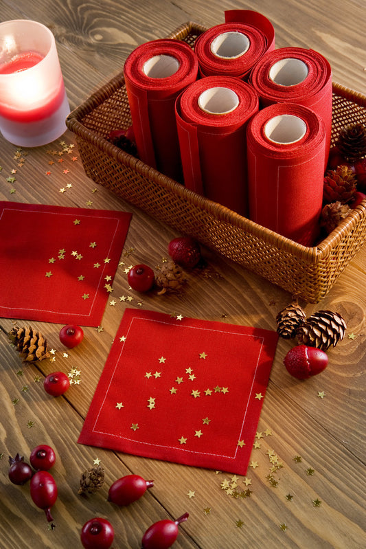 Toreador organic cotton napkin rolls in a basket with two folded Toreador organic cotton cocktail napkins on a wooden table, surrounded by star confetti, pine cones, and red berries